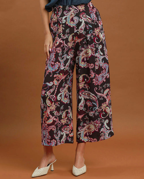 A model is shown from the waist down wearing high-waisted, wide-leg cropped pants with a vibrant paisley pattern on a black base, paired with white textured mule heels against a solid brown studio background.