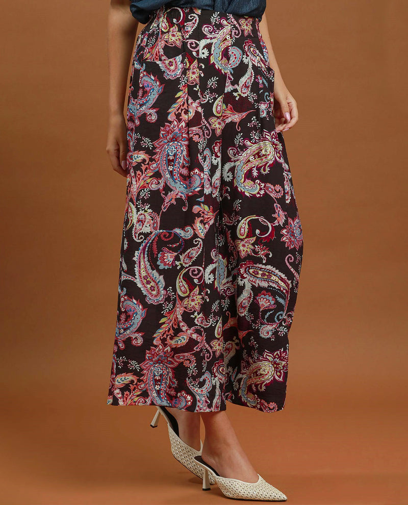 A model wearing high-waisted, wide-leg cropped pants with a vibrant paisley pattern in pink, blue, and gold on a black base, paired with white pointed-toe heels against a solid brown studio background.