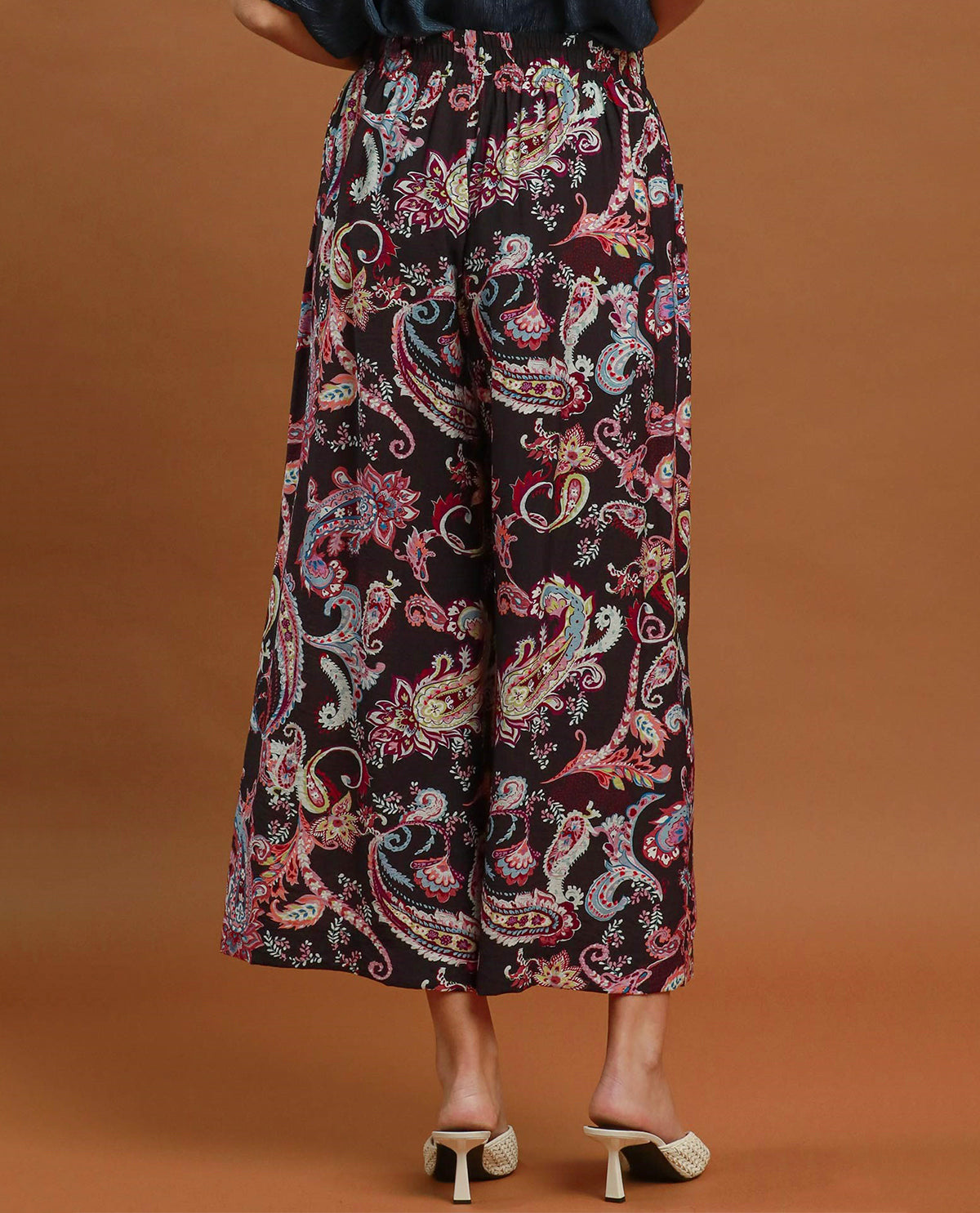 A person shown from the waist down wearing wide-leg cropped pants with a colorful paisley pattern on a black background, paired with beige heeled mule sandals against a solid brown studio backdrop.