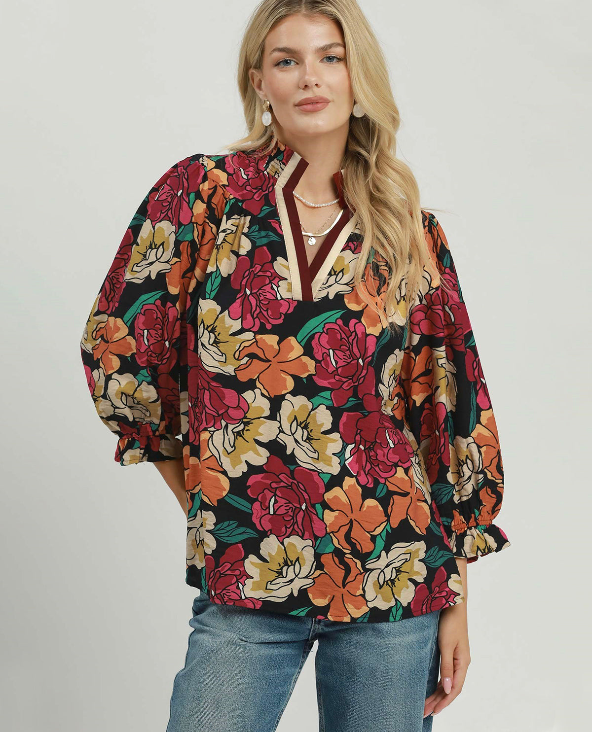 A woman wearing a black long-sleeve blouse with a vibrant multicolor floral print in red, orange, yellow, and green, paired with blue jeans, against a light gray studio background.