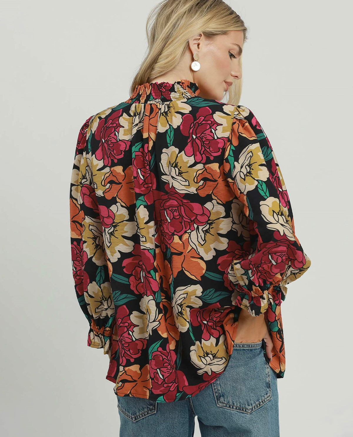 A woman wearing a black long-sleeve blouse with a vibrant multicolor floral print in red, orange, yellow, and green, paired with blue jeans, shown from a three-quarters back view against a light gray studio background.