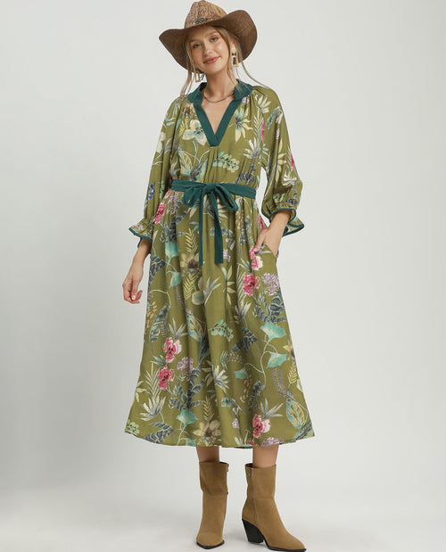 A woman modeling a long-sleeve midi dress with a green base and a colorful tropical floral print, featuring a matching green tie belt. She is also wearing a brown straw hat and tan ankle boots, standing against a plain light gray studio background.