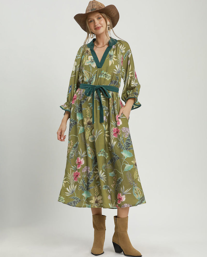 A woman modeling a long-sleeve midi dress with a green base and a colorful tropical floral print, featuring a matching green tie belt. She is also wearing a brown straw hat and tan ankle boots, standing against a plain light gray studio background.