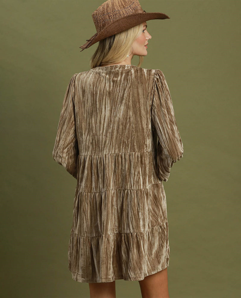 A woman wearing a brown tiered velvet dress with a textured, crinkled finish and a matching brown wide-brim hat, shown from the back against an olive green background.