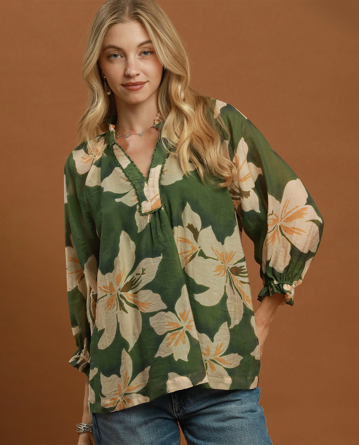 A woman wearing a green floral blouse with large cream and peach flowers, paired with blue jeans, standing against a solid brown studio background.