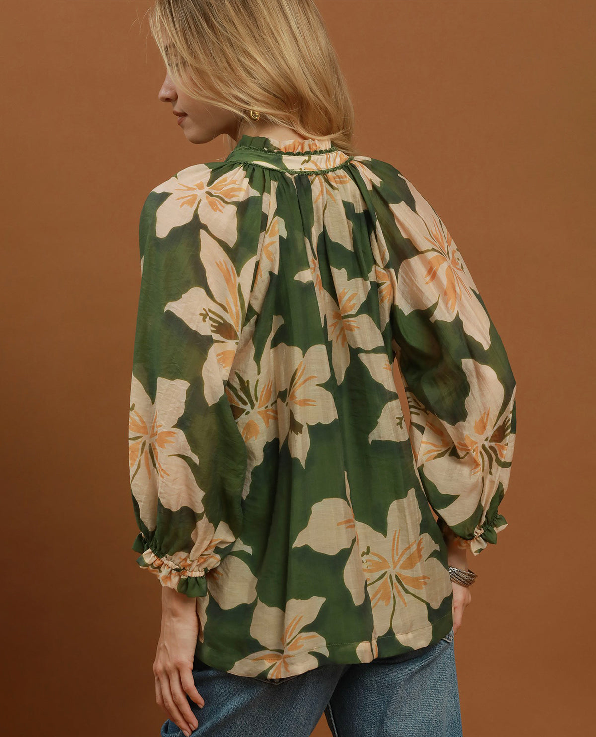 A woman shown from the back wearing a green long-sleeve blouse with a large cream floral print and blue jeans, standing against a solid warm brown background.
