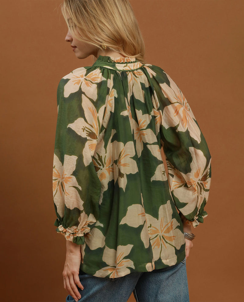 A woman shown from the back wearing a green long-sleeve blouse with a large cream floral print and blue jeans, standing against a solid warm brown background.