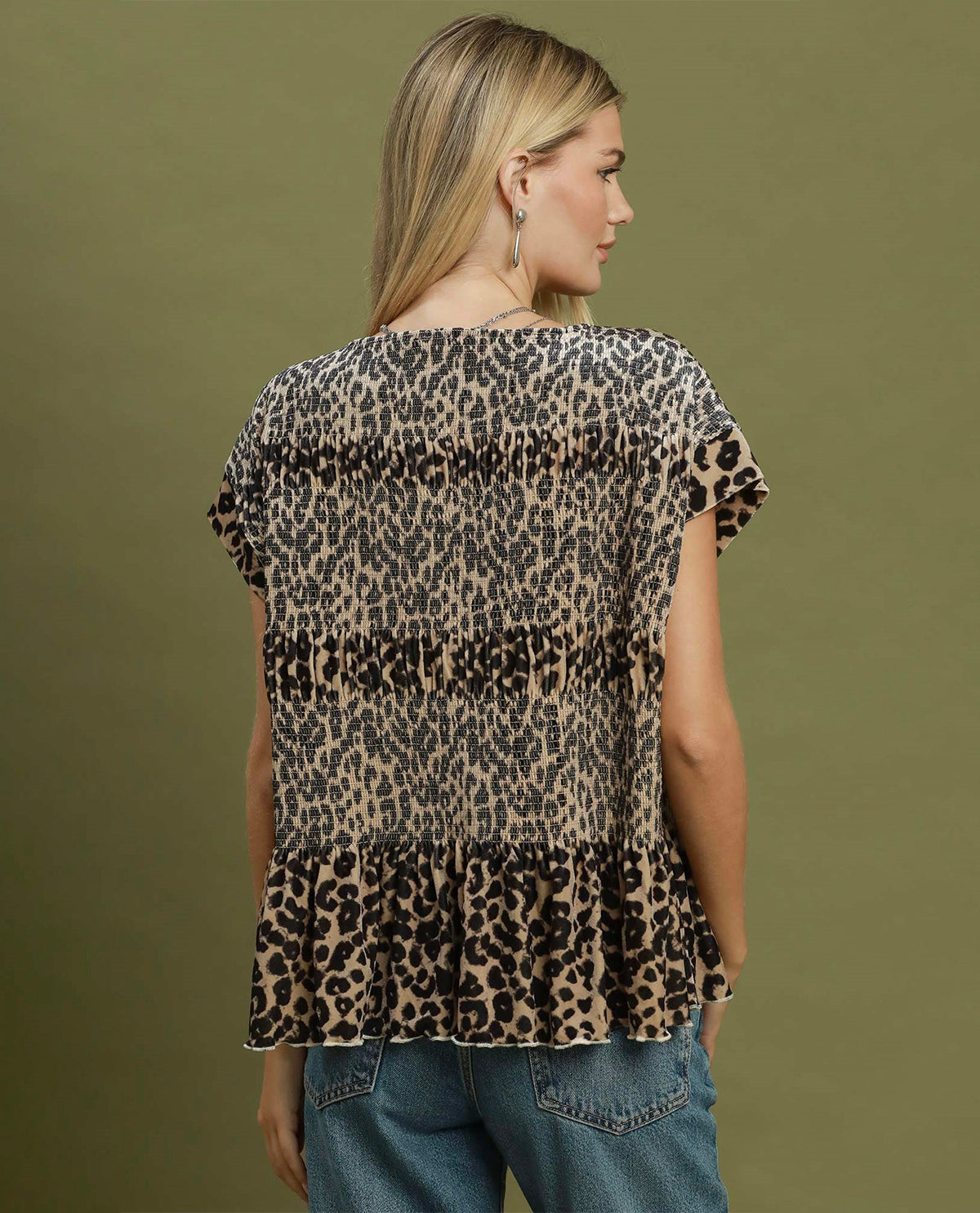 A woman shown from the back wearing a leopard-print short-sleeve blouse with a ruffled hem, paired with blue jeans, against a solid olive green background.