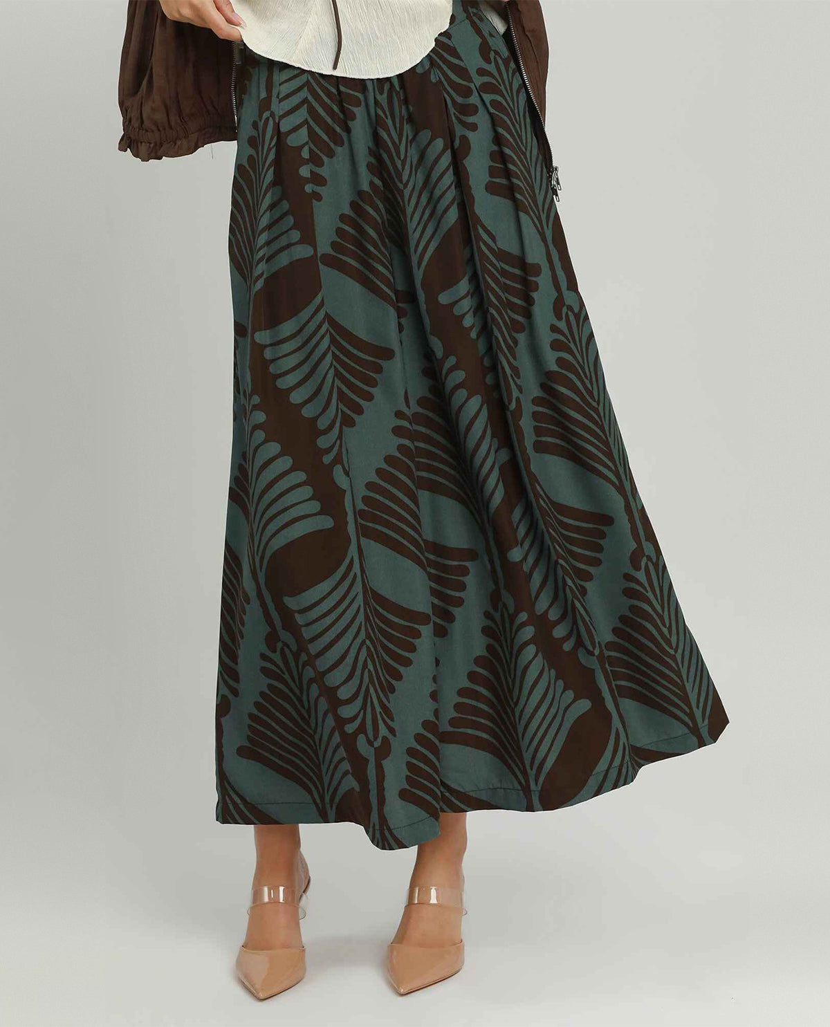 A woman models a long, wide-leg pants featuring a dark brown and muted teal-green fern leaf pattern, styled with a cream top and beige heels against a light gray studio background.