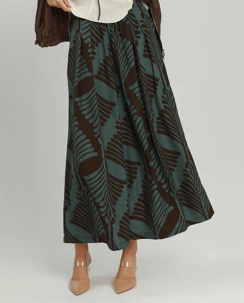A woman models a long, wide-leg pants featuring a dark brown and muted teal-green fern leaf pattern, styled with a cream top and beige heels against a light gray studio background.