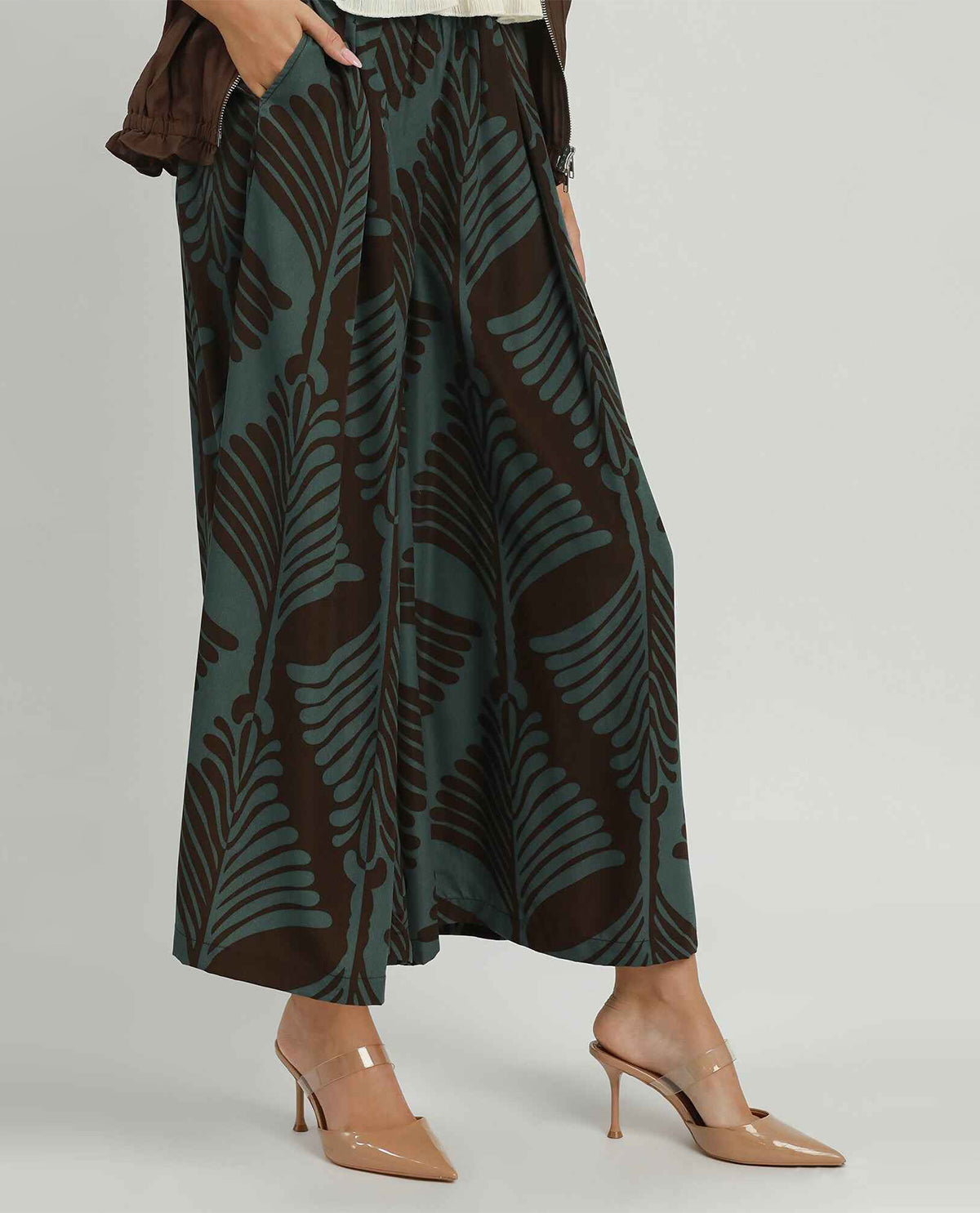 A model wearing wide-leg cropped pants with a dark brown and teal palm leaf pattern, paired with beige pointed-toe mule heels against a light gray studio background.
