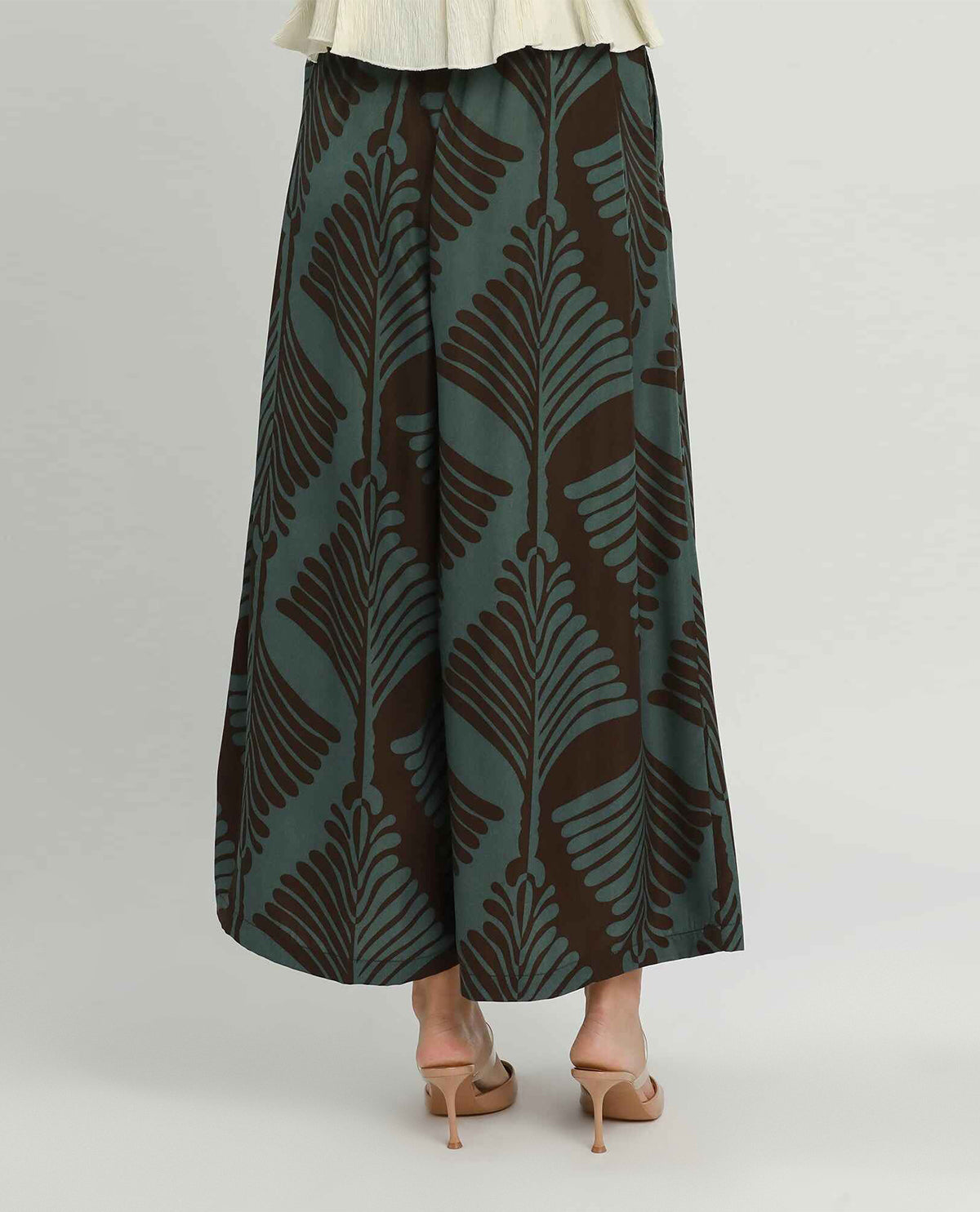 A person shown from the waist down wearing wide leg pants with a dark brown and teal palm leaf pattern, paired with beige high-heeled sandals against a light gray studio background.
