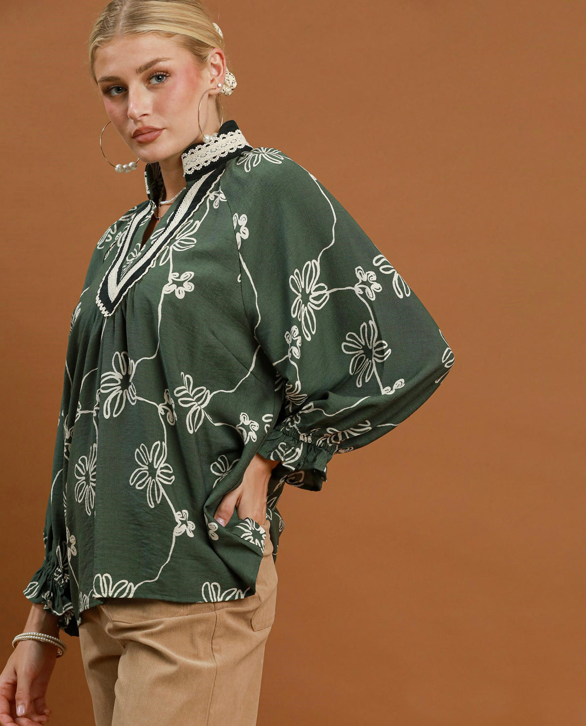 A woman wearing a dark green long-sleeve blouse with a white floral vine pattern, paired with beige trousers, posed against a solid warm brown background.