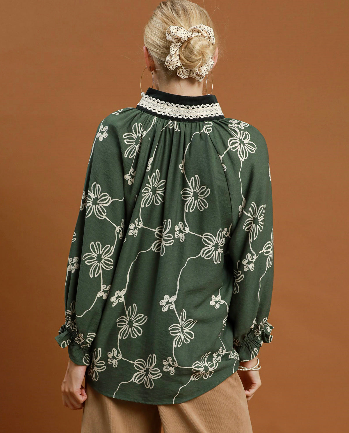 A woman viewed from behind wearing a dark green long-sleeve blouse with a white floral pattern, a black collar with white lace trim, and beige pants, set against a solid warm brown background.