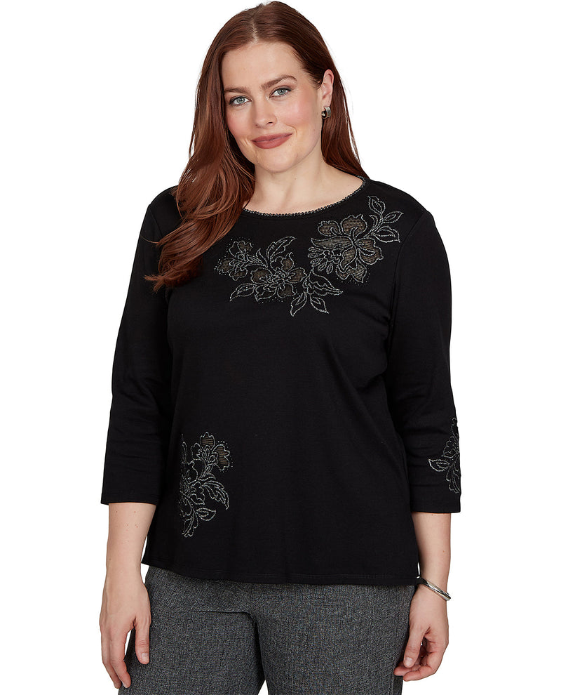 A woman wearing a black three-quarter sleeve top with silver floral embroidery on the chest and sleeve, paired with gray pants, against a plain white background.