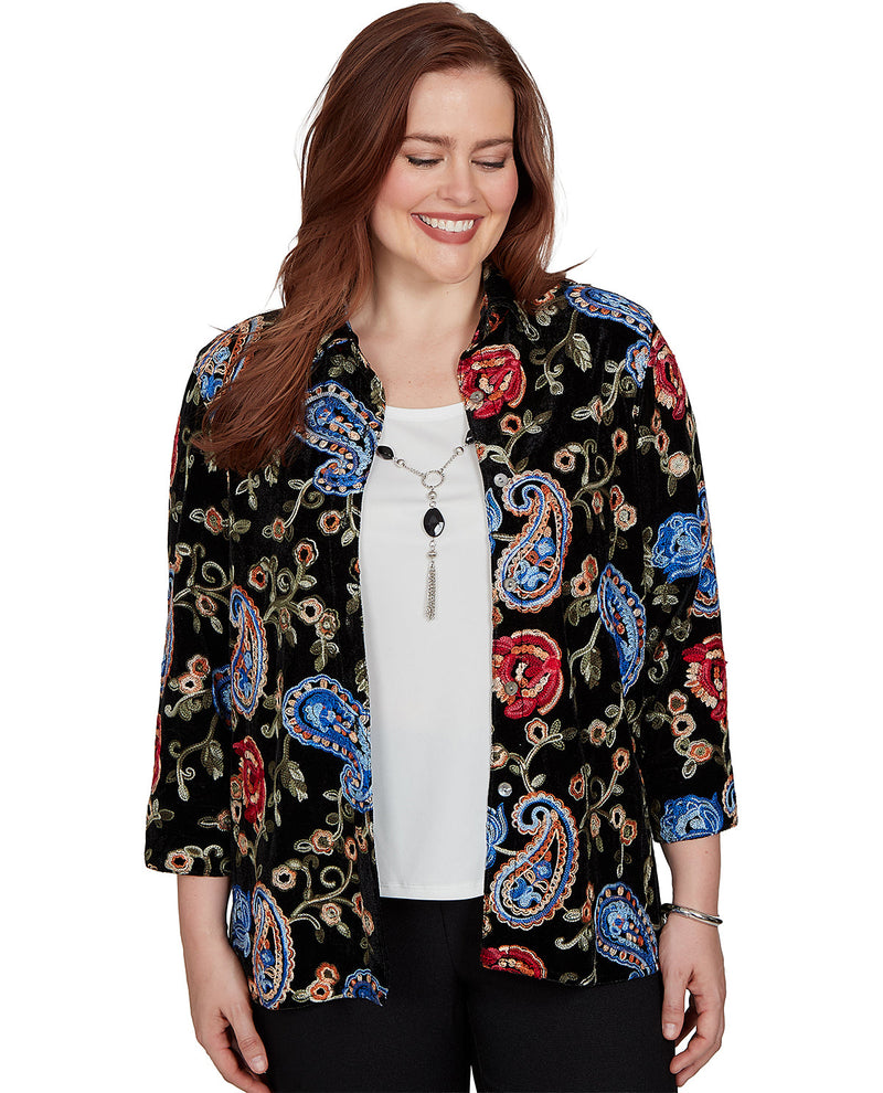 A woman wearing a black button-up shirt with a colorful paisley and floral pattern in red, blue, beige, and green, layered over a white top and black pants, against a plain white background.