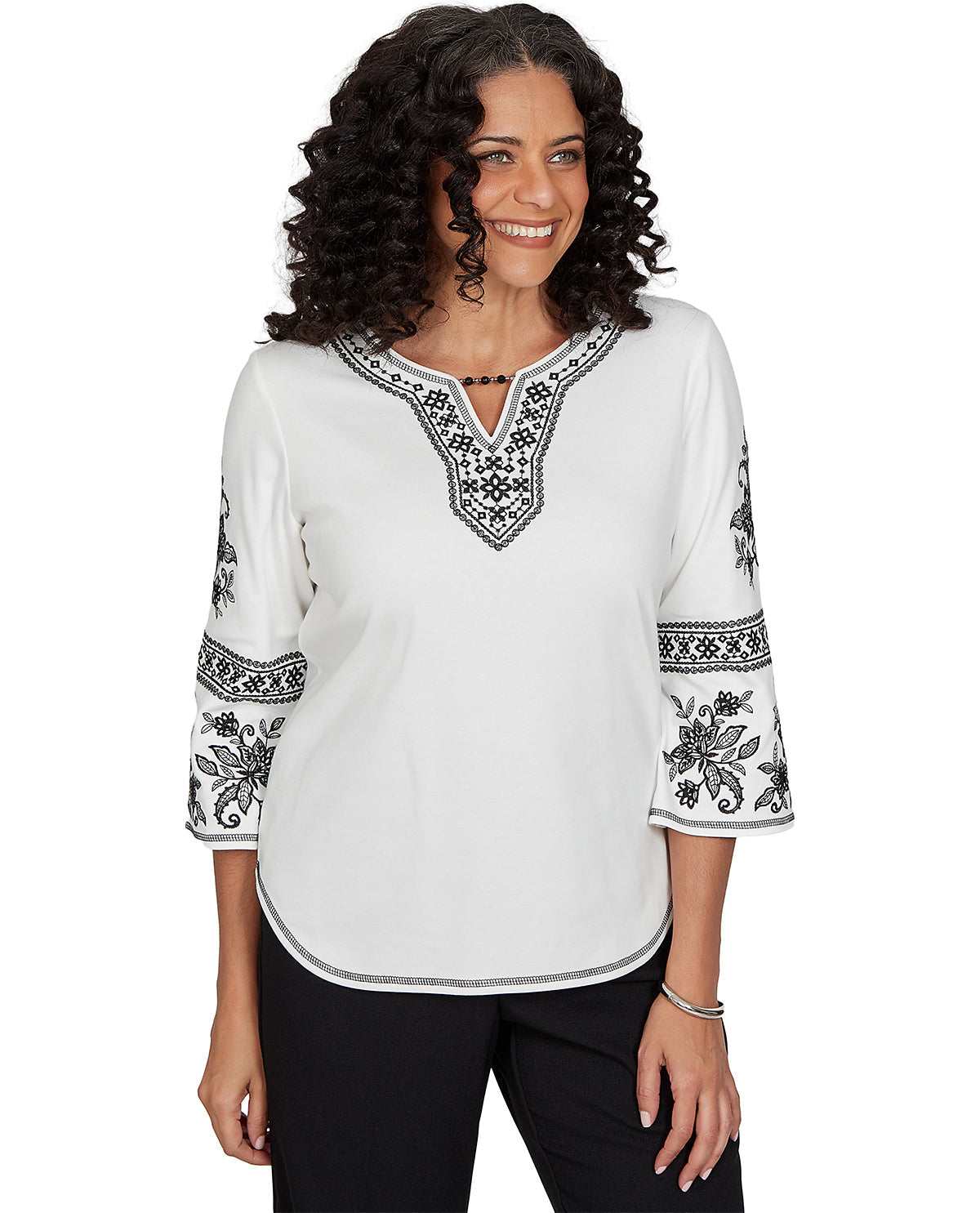 A woman wearing a white three-quarter sleeve top with black geometric and floral embroidery, paired with black pants, against a plain white background.