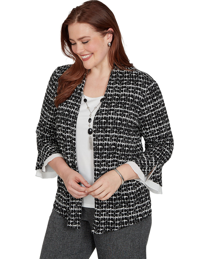A woman wearing a black and white tweed open-front jacket with flared three-quarter sleeves, layered over a white top and gray pants, photographed against a plain white background.