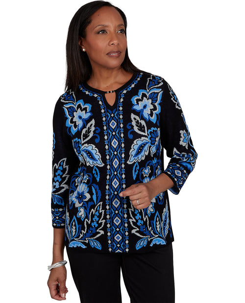 A woman modeling a black long-sleeve top with vibrant blue and white floral and geometric patterns, paired with black pants against a plain white background.