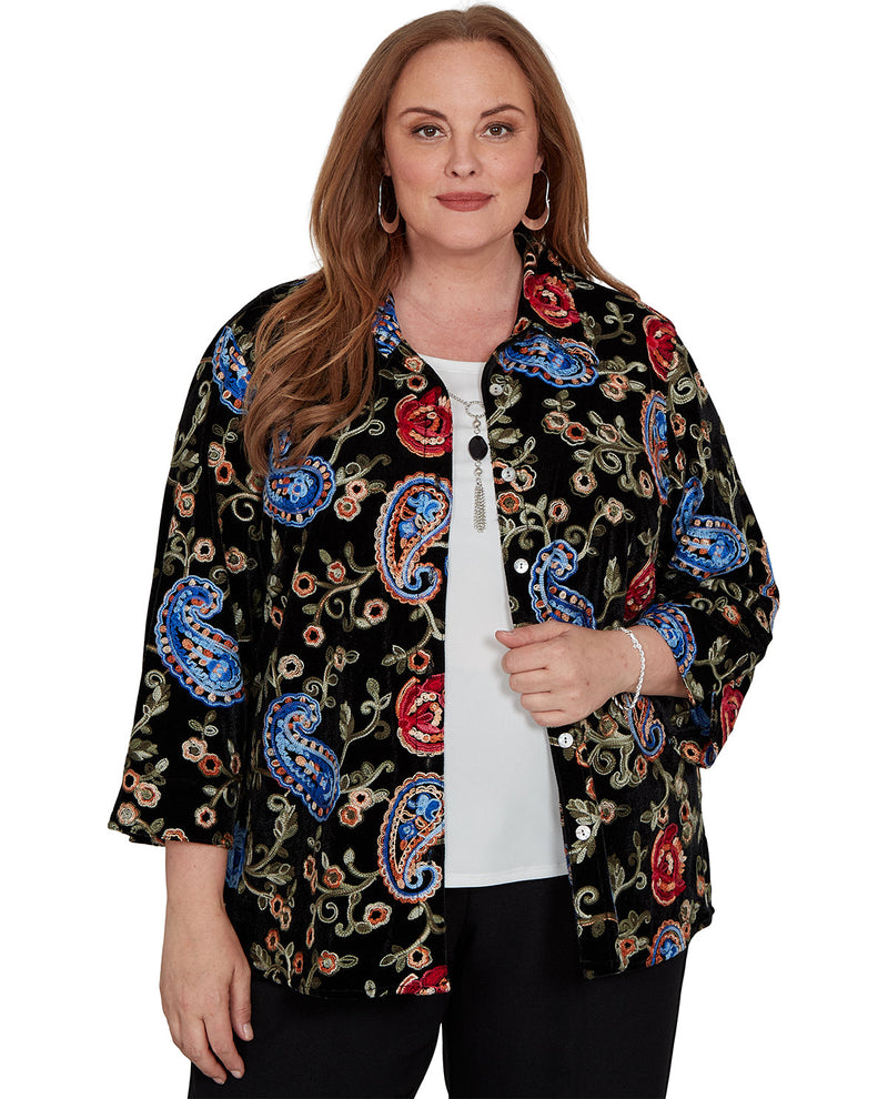 A woman modeling a black button-up shirt with a vibrant multicolor paisley and floral pattern, worn open over a white top and black pants, against a plain white background.