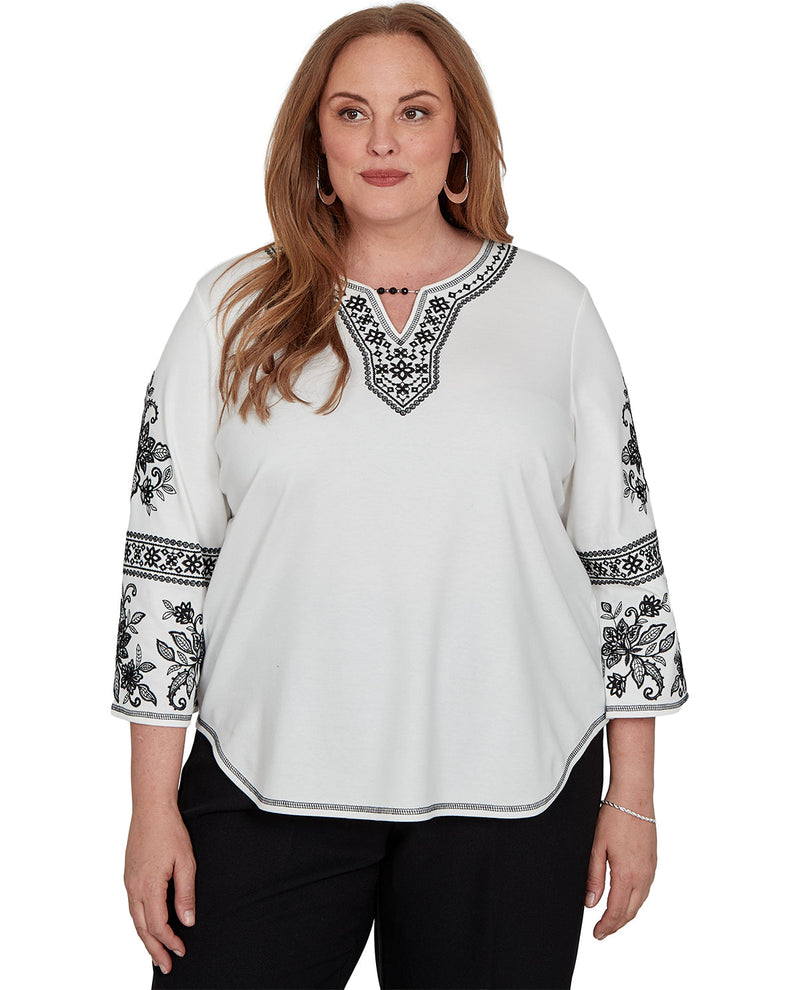 A woman modeling a white three-quarter sleeve blouse with black floral and geometric embroidery on the neckline and sleeves, paired with black pants against a plain white background.