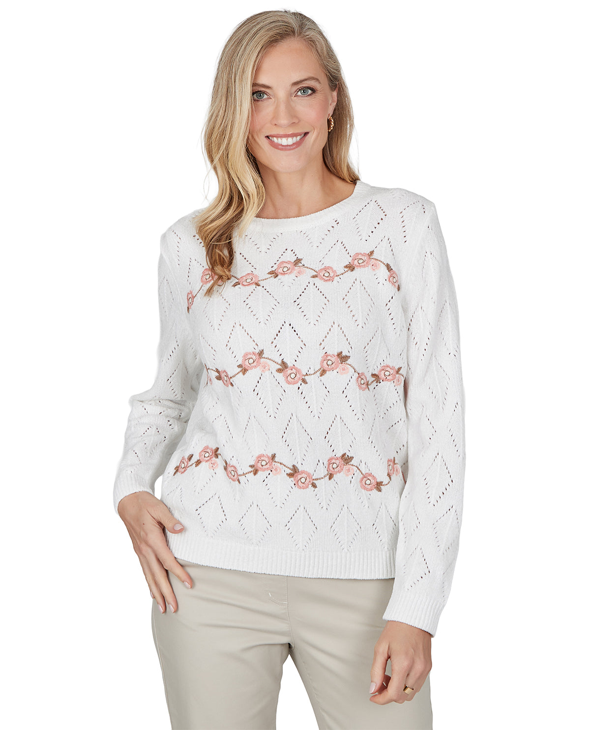 A woman modeling a white long-sleeve knit sweater with a diamond lattice pattern and horizontal rows of pink floral embroidery, paired with light beige pants against a plain white background.