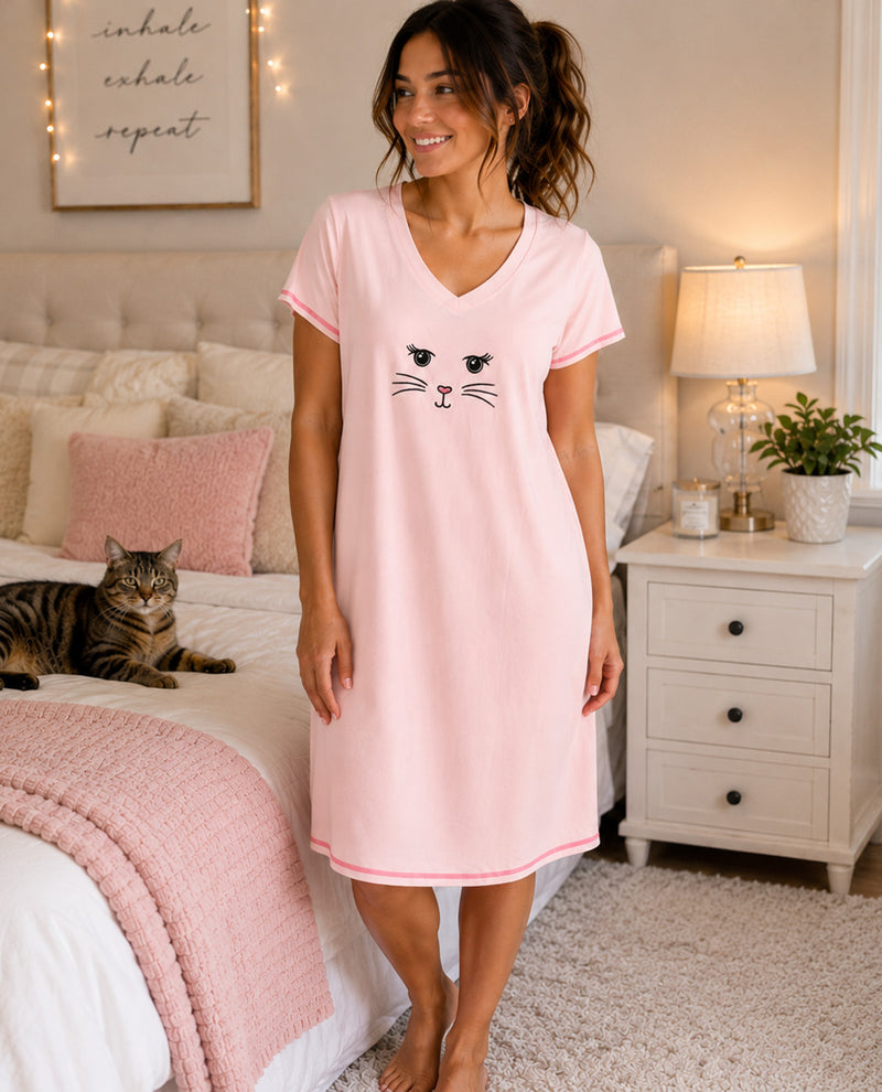 A woman wearing a light pink short-sleeve V-neck nightshirt with a black cat face graphic on the front, standing in a cozy bedroom next to a bed with a cat lying on it.