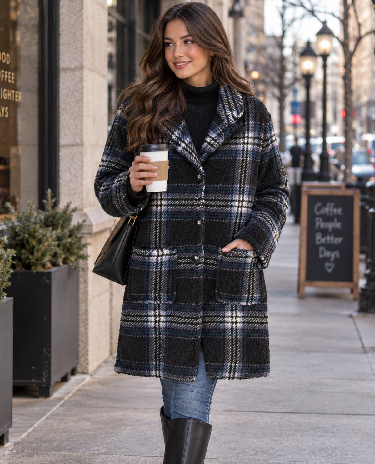 A woman stands on a city sidewalk wearing a long black, white, and blue plaid coat over a black turtleneck, blue jeans, and black boots, while holding a coffee cup and carrying a black handbag.
