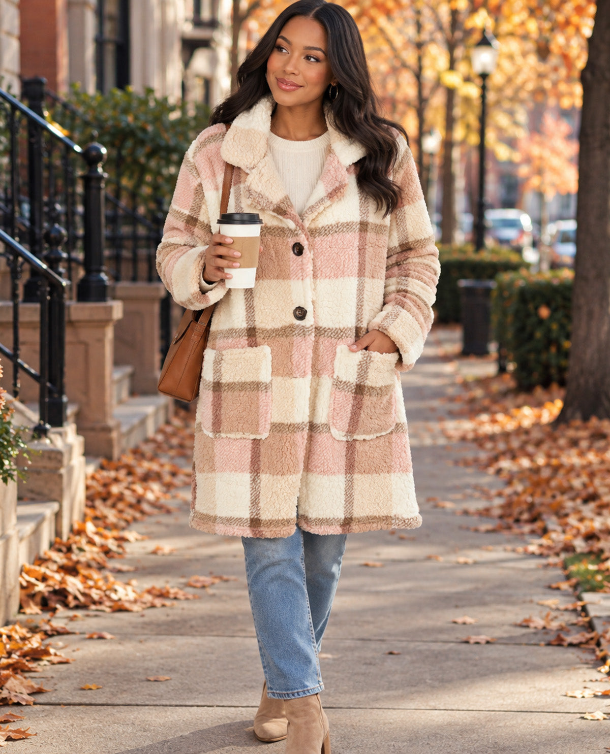 A woman walking on an autumn city sidewalk wearing a plush beige and blush pink plaid coat over a cream sweater, blue jeans, and tan ankle boots, while holding a coffee cup and carrying a brown shoulder bag.