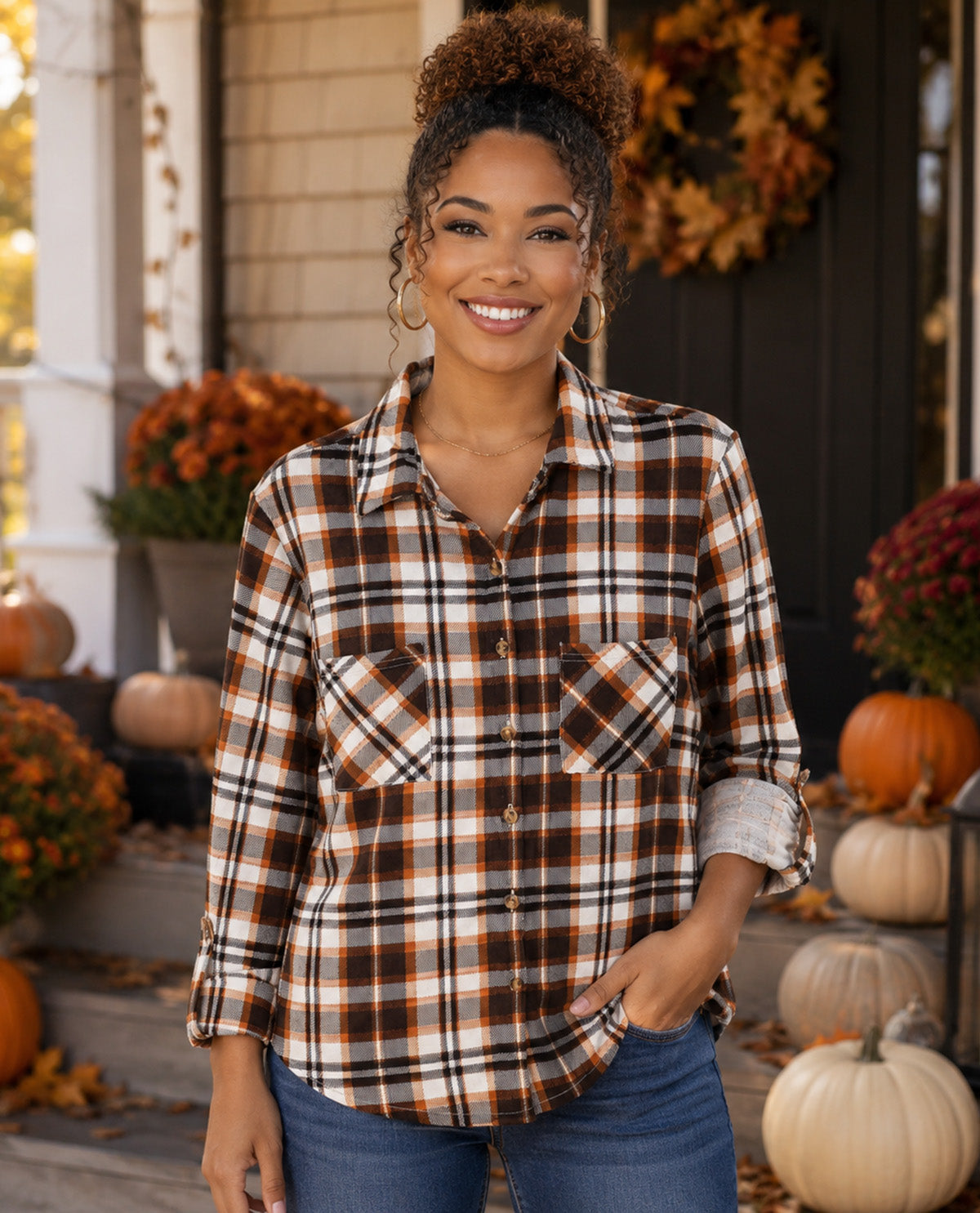 A woman stands on a decorated front porch wearing a brown, orange, and white plaid button-up shirt with blue jeans. Pumpkins, fall flowers, and a wreath are visible in the background.