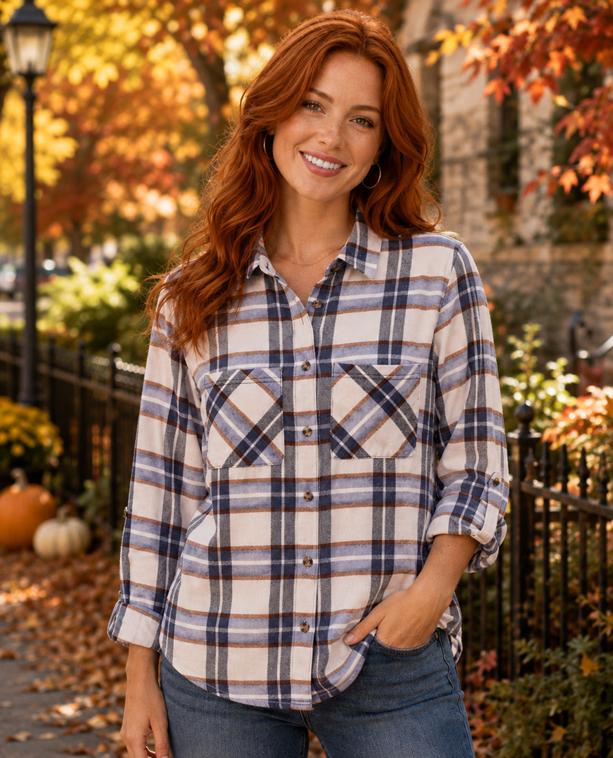 A smiling woman with long red hair stands outdoors wearing a long-sleeve plaid button-up shirt in cream, navy, and brown tones, paired with blue jeans. The background shows autumn trees with orange and yellow leaves, a black fence, and pumpkins.