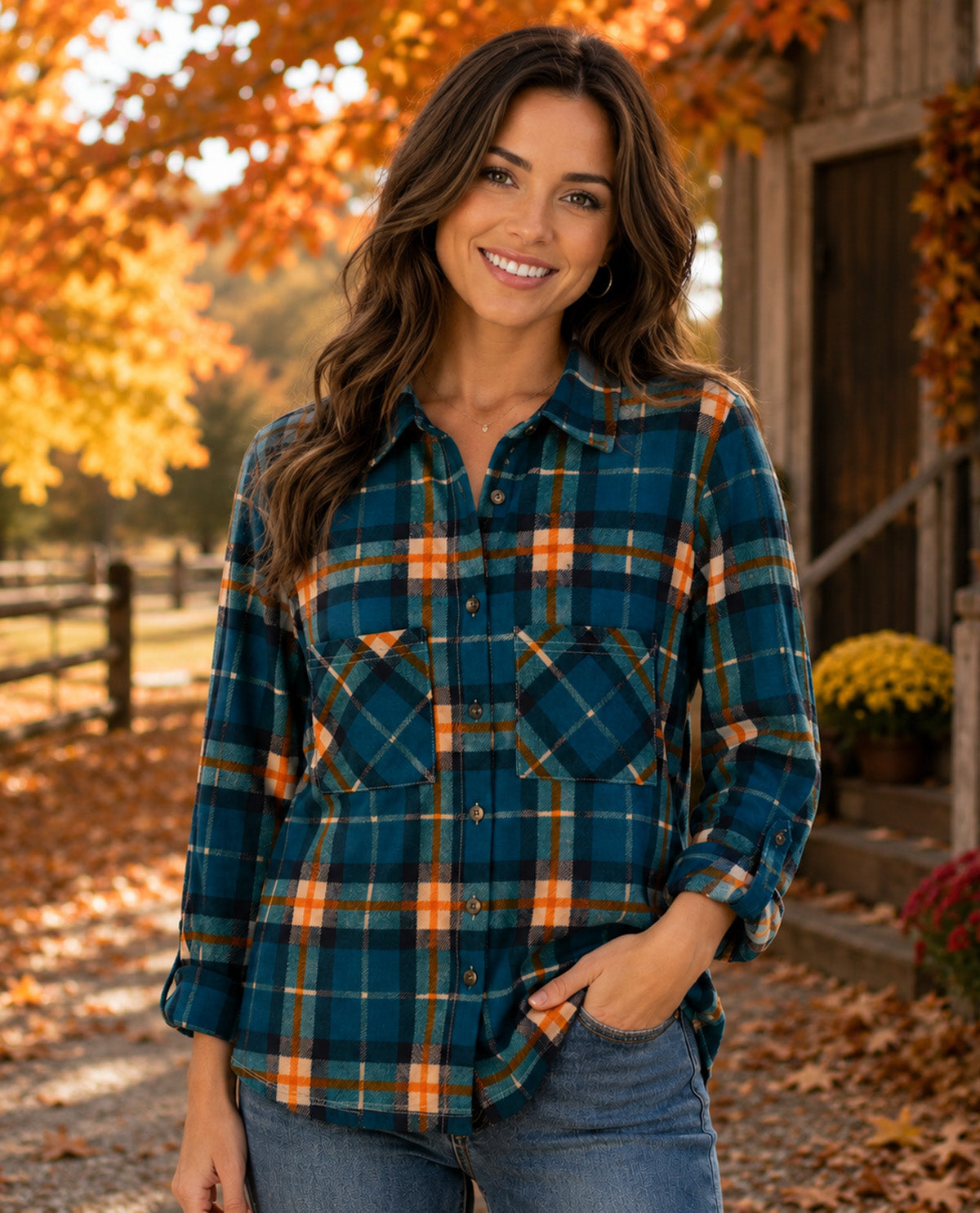 A woman wearing a teal, orange, and brown plaid button-down flannel shirt, standing outdoors in an autumn setting with fallen leaves and a rustic wooden cabin in the background.