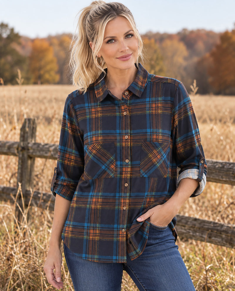 A woman wearing a dark navy plaid button-up shirt with blue and orange stripes, standing in a dry grass field with a wooden fence and autumn trees in the background.