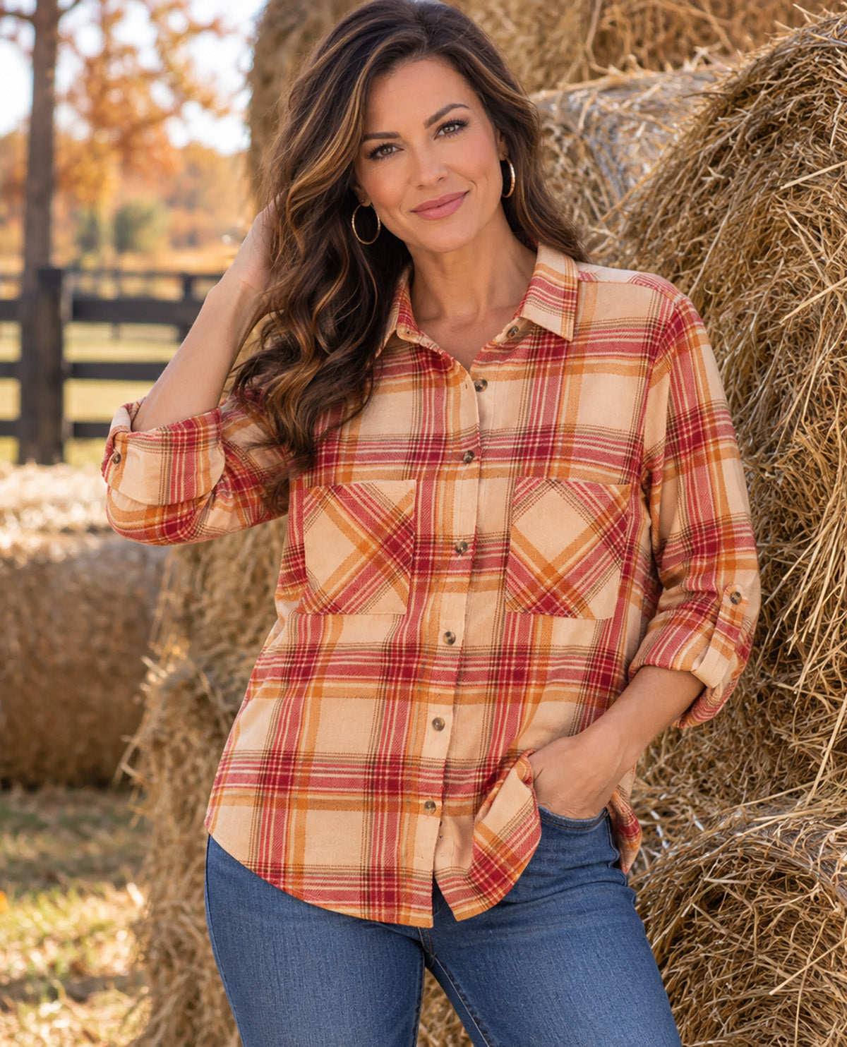 A woman wearing a long-sleeve plaid button-up shirt in beige, orange, and red tones, paired with blue jeans, standing outdoors in front of hay bales.