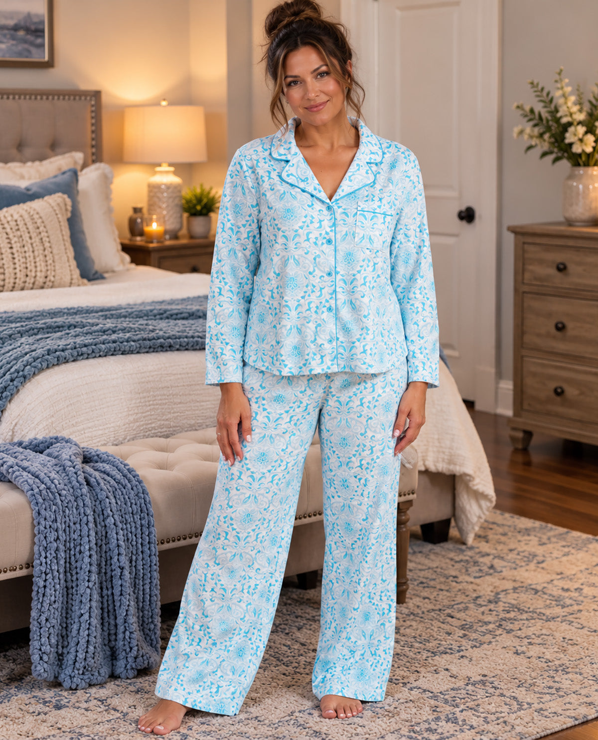 A woman stands barefoot in a bedroom wearing a light blue pajama set with a white floral print. The set includes a long-sleeve button-front top and matching wide-leg pants.