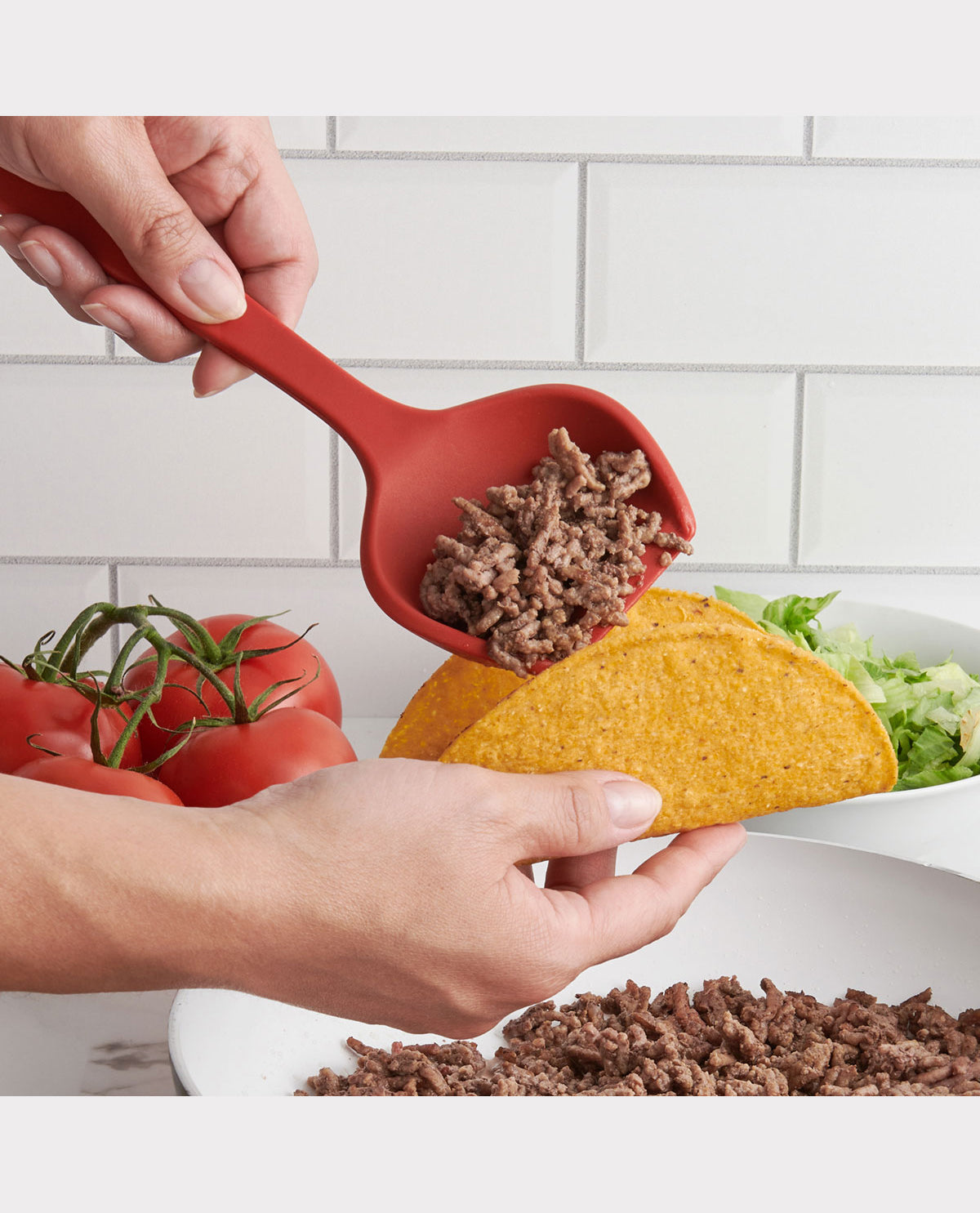 Person adding ground beef to a taco with a red spoon, surrounded by tomatoes and lettuce on a white surface.