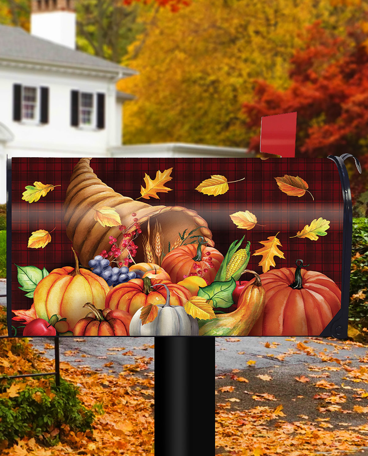 A decorative mailbox cover featuring a Thanksgiving harvest scene with pumpkins, corn, grapes, leaves, and a cornucopia on a red plaid background, shown installed on a mailbox in an autumn yard with trees and fallen leaves visible.