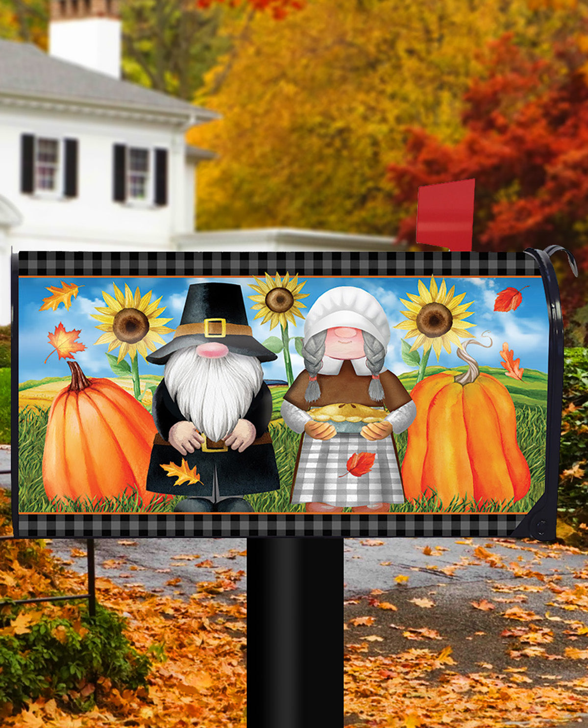 A decorative mailbox covered in an autumn-themed printed design featuring a pilgrim and a woman with sunflowers, pumpkins, leaves, and a black-and-white checkered top border. The mailbox stands outdoors on a post with a blurred fall yard background of trees, leaves, and a house.