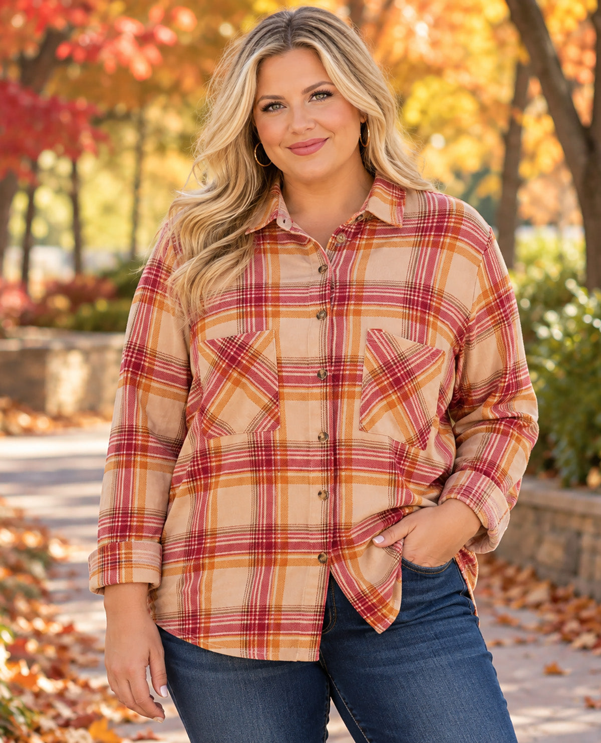 A woman wearing a long-sleeve plaid button-up shirt in beige, orange, and red tones, paired with blue jeans, standing outdoors on a path with autumn trees in the background.