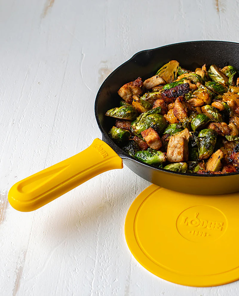 A black cast iron skillet with a bright yellow handle and matching yellow lid, shown on a white textured surface with cooked Brussels sprouts and chicken inside the pan.