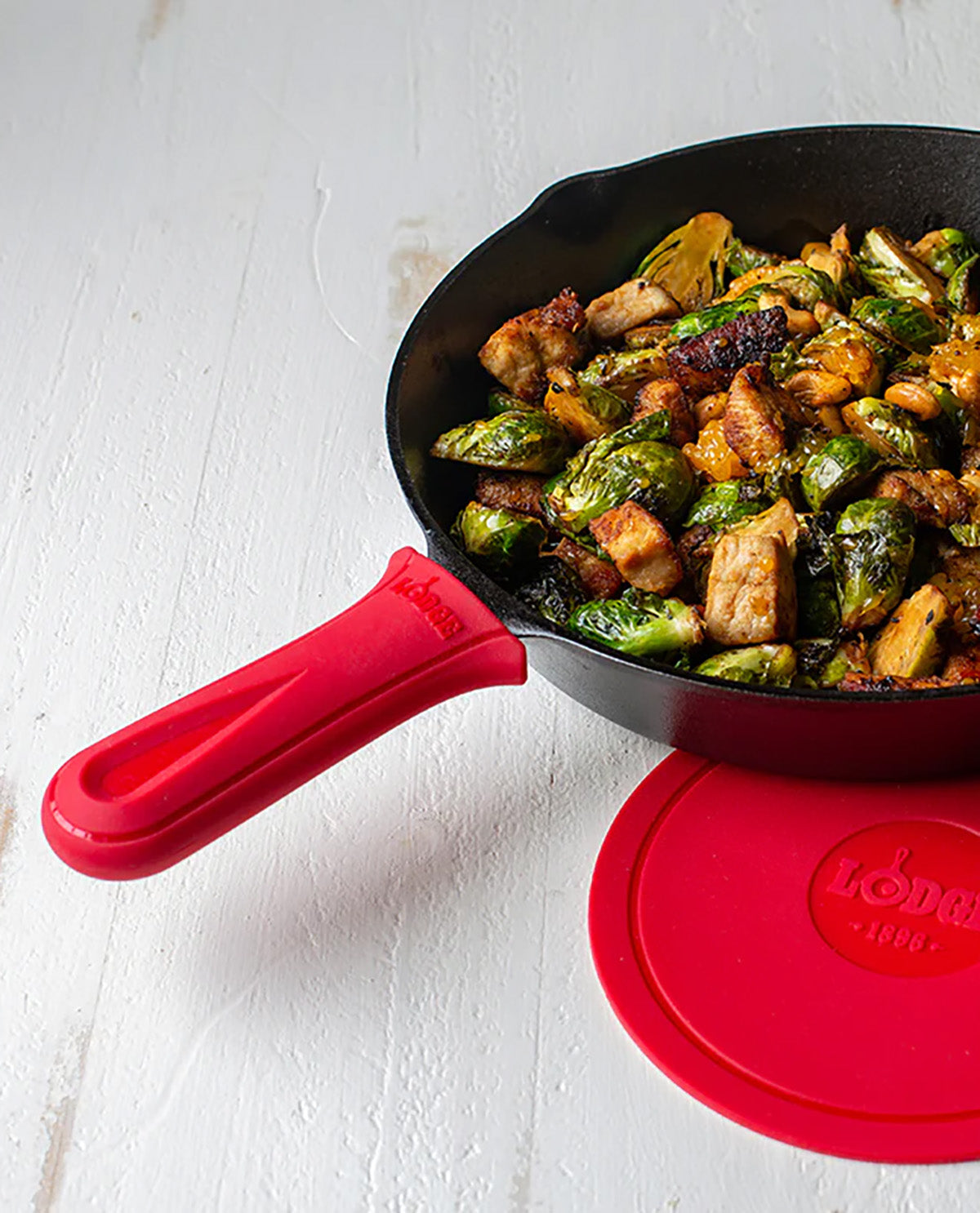 A black cast iron skillet with a bright red silicone handle and matching red lid, shown on a white textured surface with cooked food inside the pan.