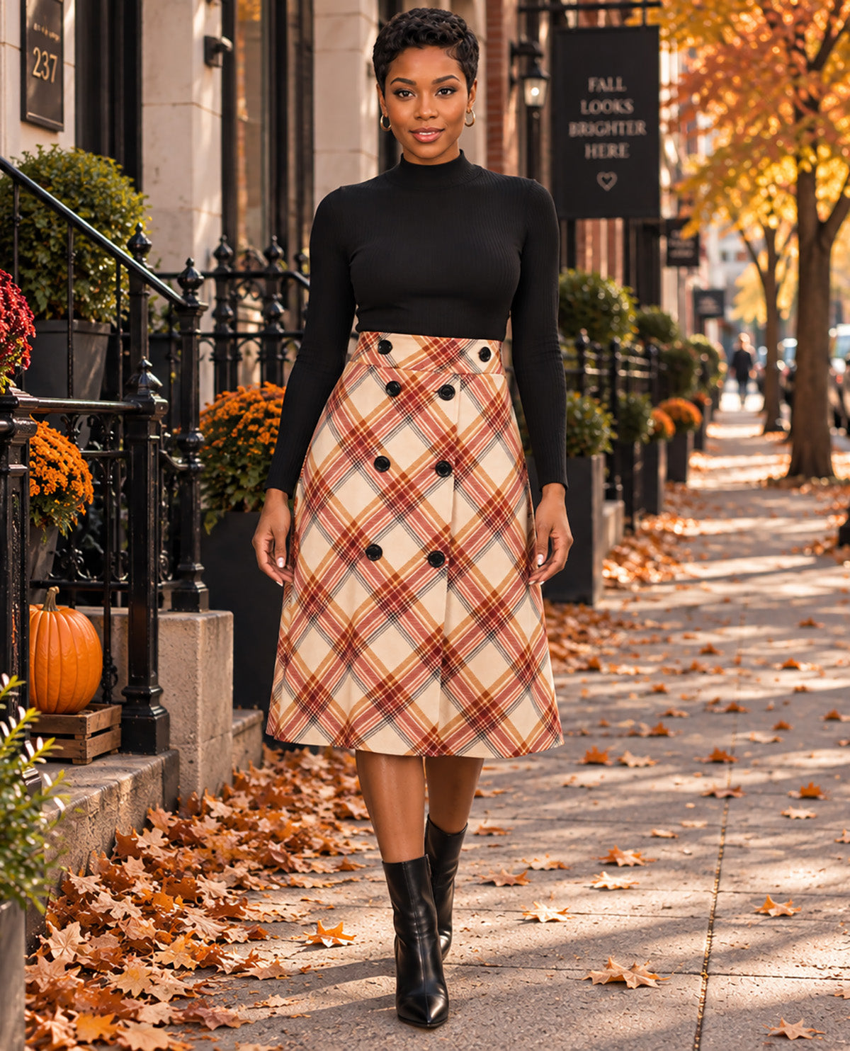 A woman stands on an autumn city sidewalk wearing a black long-sleeve mock-neck top and a knee-length plaid skirt with black ankle boots. The sidewalk is scattered with fallen leaves, and the background shows a building entrance with potted plants, a pumpkin, and fall foliage.