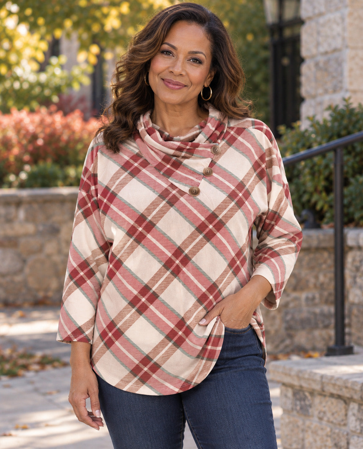A woman wearing a long-sleeve cowl-neck plaid top in beige and rust red, paired with dark blue jeans, standing outdoors in front of a stone building and greenery.