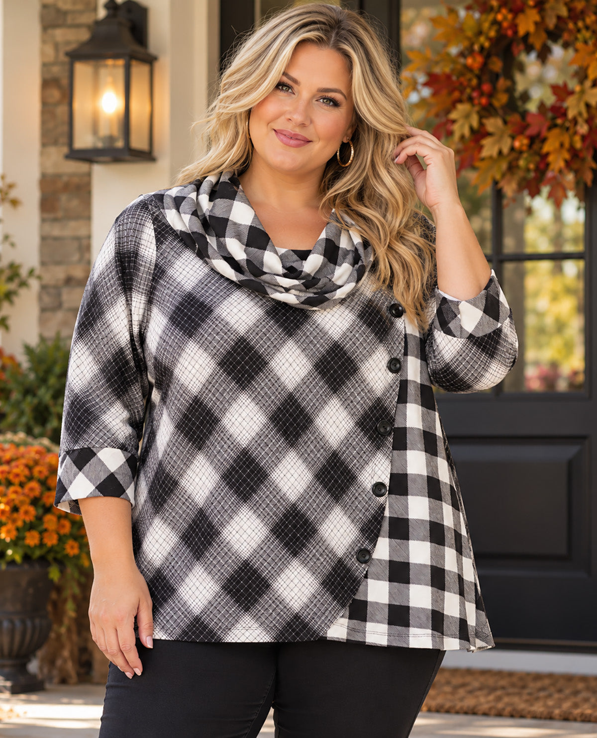 A woman models a black and white buffalo plaid wrap-style top with three-quarter sleeves and a button-front overlay, paired with black pants, set against a warm autumnal front porch background.