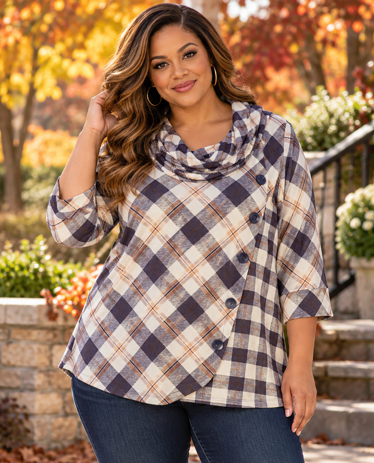 A woman wearing a plaid cowl-neck top with three-quarter sleeves and dark buttons, standing outdoors in front of a stone staircase and autumn garden.