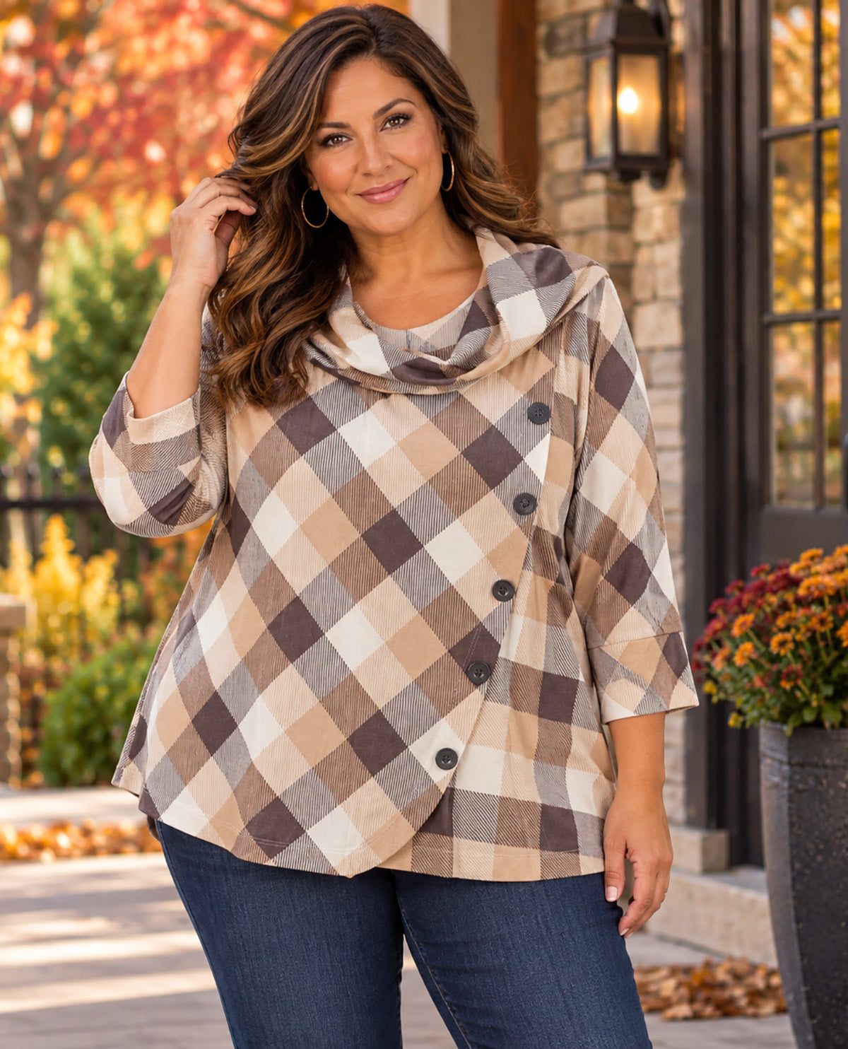 A woman wearing a beige, brown, and white plaid wrap-style top with a cowl neck and dark buttons, paired with blue jeans, standing outdoors in front of a stone wall and autumn foliage.