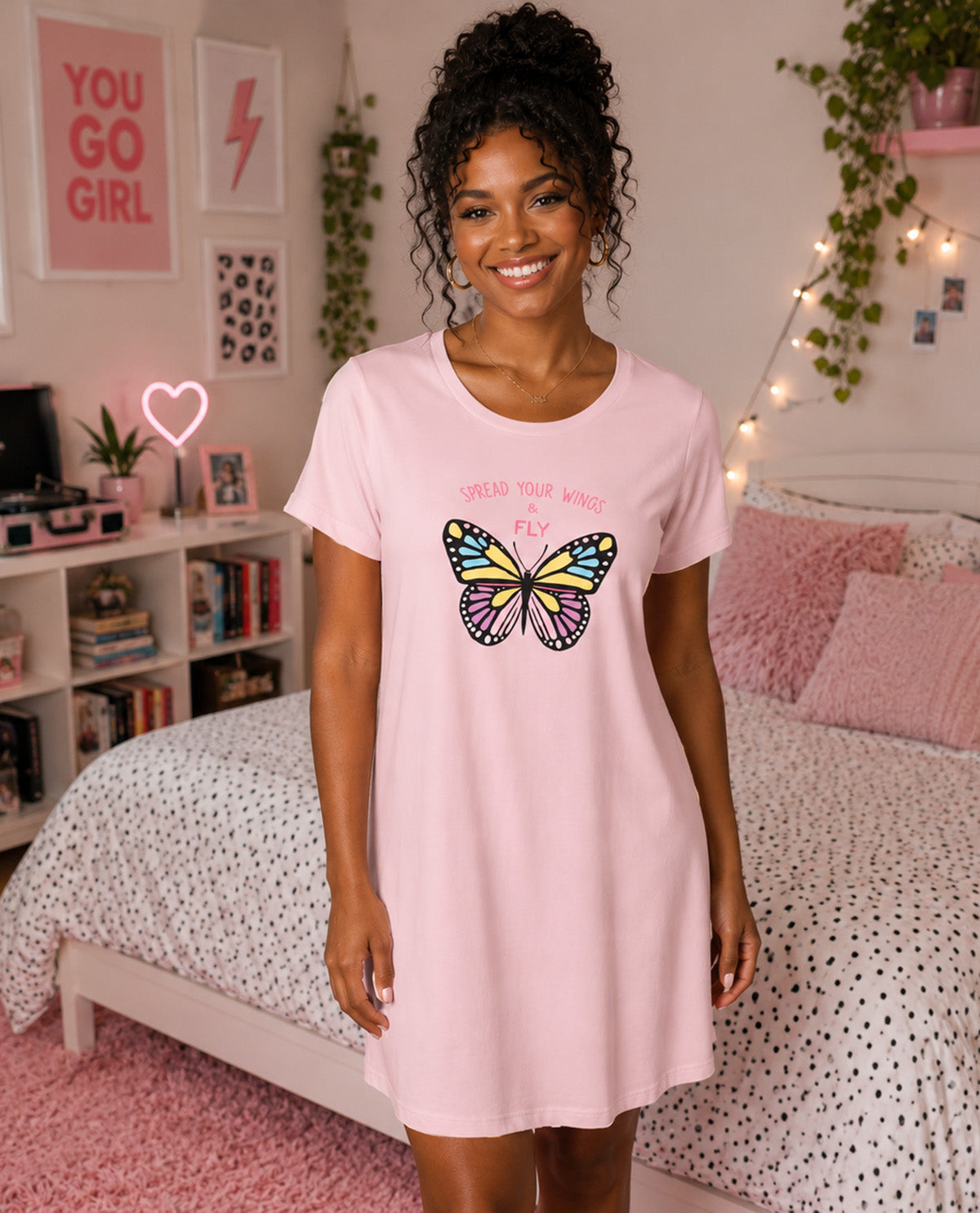 A woman wearing a light pink t-shirt nightdress with a colorful butterfly graphic and the text 'SPREAD YOUR WINGS AND FLY' stands in a pink-themed bedroom.