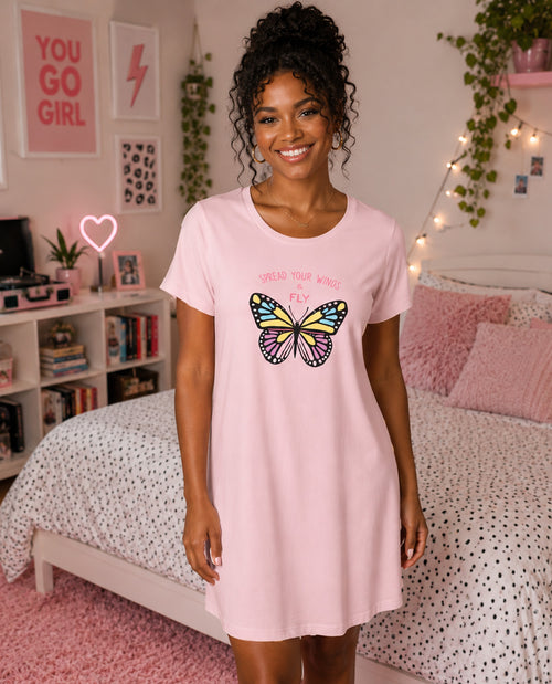 A woman wearing a light pink t-shirt nightdress with a colorful butterfly graphic and the text 'SPREAD YOUR WINGS AND FLY' stands in a pink-themed bedroom.