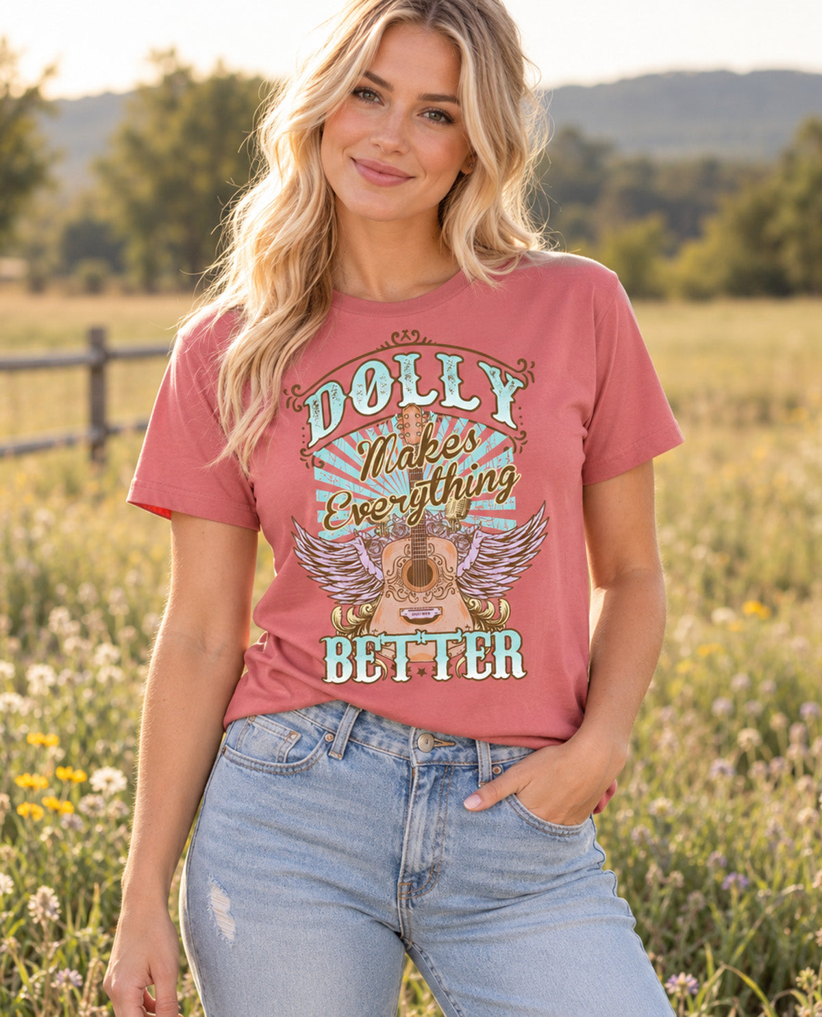 A woman wearing a dusty rose graphic t-shirt with a western 'Dolly Makes Everything Better' design, paired with light-wash blue jeans, standing in a sunny, grassy field.