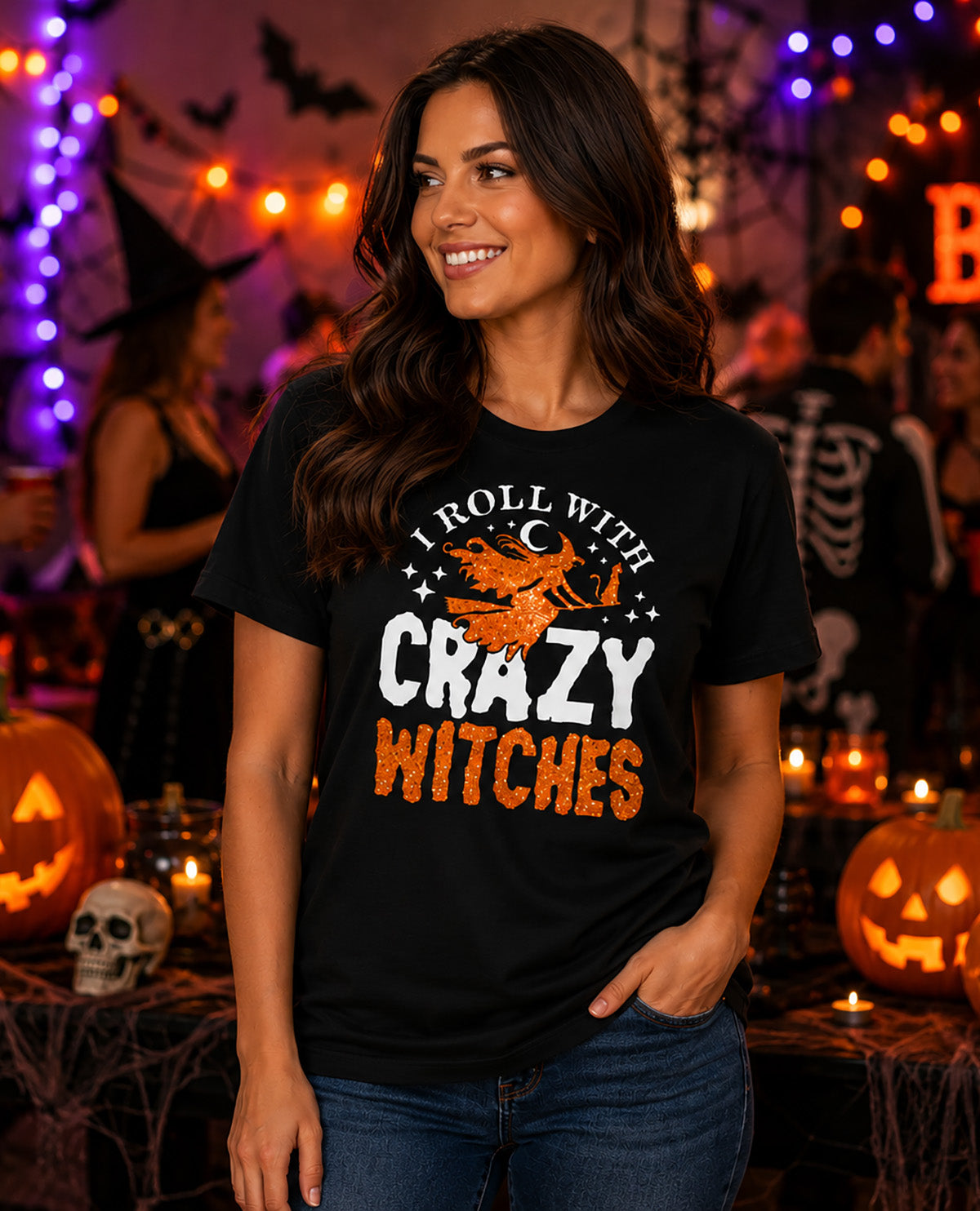 A woman wearing a black graphic t-shirt with the text 'I ROLL WITH CRAZY WITCHES' and an illustration of a witch riding a broomstick, set against a Halloween-themed background with pumpkins, lights, and other partygoers.